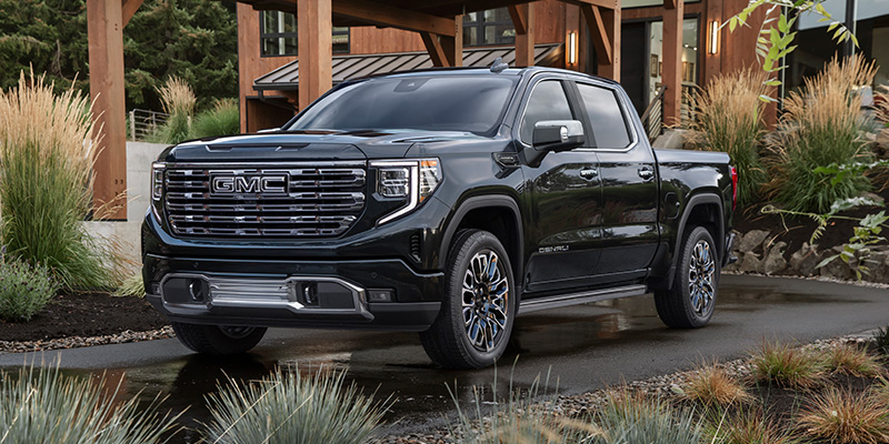 Front-three-quarter view of a black GMC Sierra Denali pickup truck parked on a paved driveway outside a modern wooden home.
