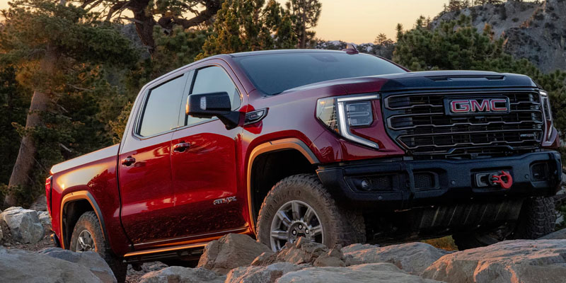 Front-three-quarter view of a dark red GMC Sierra AT4X pickup truck driving over rocky off-road terrain in a mountainous forest setting.