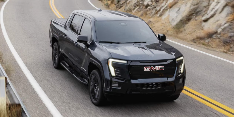 High-angle front-three-quarter view of a dark gray GMC Sierra EV electric pickup truck driving along a curved, paved road next to a rocky hillside.